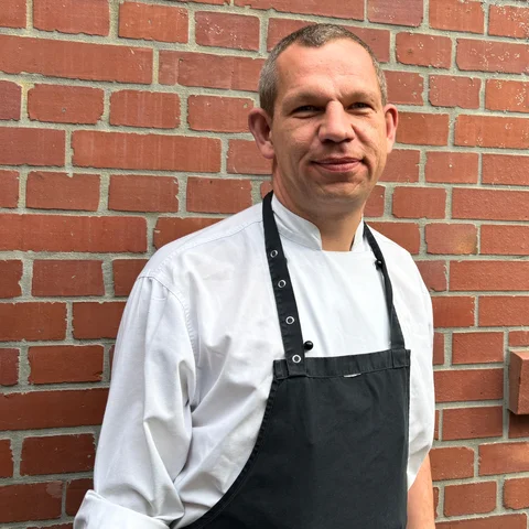 Matthias Hartmann der pädagogische Leiter der SozDia Inklusionsküchen vor eiiner Backsteinwand mit weißer Kochkleidung.