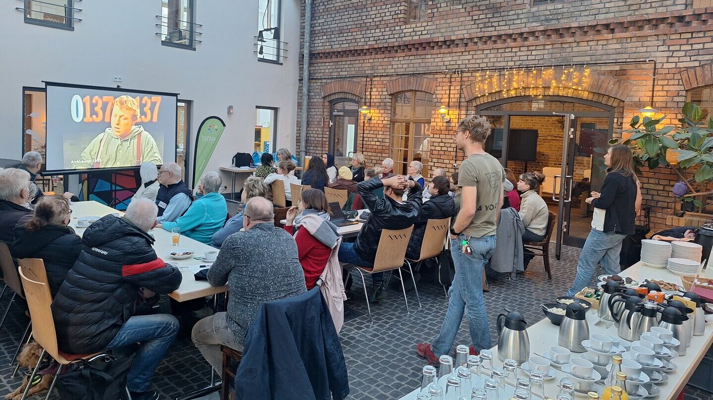 Eine Gruppe Freiwilliger sitzt án einem Tisch und schaut einen Film. Es gibt ein Buffet und der Raum ist hell.