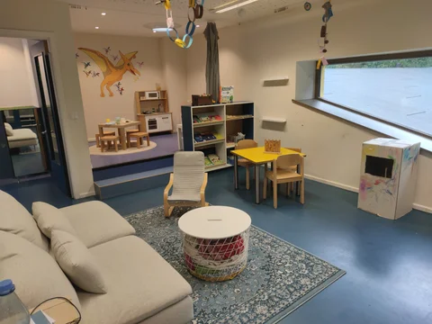 Ein farbenfroher, kinderfreundlicher Raum mit Sofa, Spielbereichen wie Küche, kleinen Tischen, Bücherregal und Pterodaktylus-Wandbild.