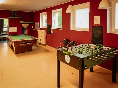Ein Spielzimmer mit roten Wänden, Billardtisch, Kickertisch und offener Tür zu einem weiteren Loungebereich.