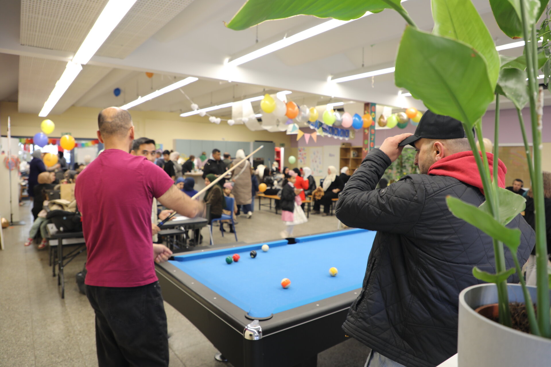 Männer spielen Billard bei einem lebhaften Gemeinschaftsereignis. Viele Leute unterhalten sich an Tischen, bunte Ballons schmücken den Raum.