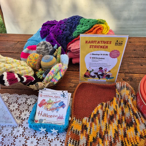 Tisch mit handgestrickten und gehäkelten Artikeln in verschiedenen Farben, einem Schild für "Karitatives Stricken" und Handwerkskarten.