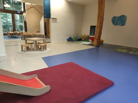 Ein geräumiger Kinderspielbereich mit Essplatz, Entspannungsecke, Rutsche und Spielmatten in einem hellen Raum.