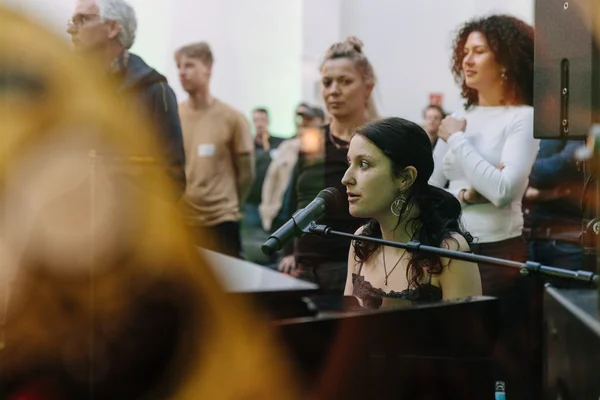 Eine junge Frau sitzt am Klavier und singt. Hinter stehen Menschen.