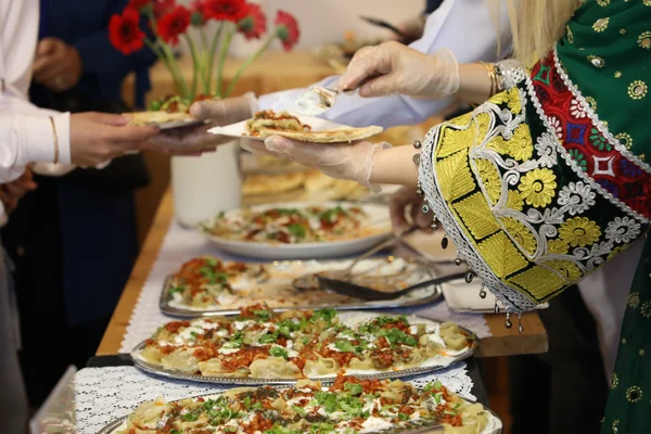 Person in bestickter traditioneller Kleidung serviert Teigtaschen von einem Buffet an Gäste.