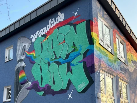 Eine Hausfassade mit einem großflächigen, farbenfrohen Graffiti. Im Zentrum steht das Wort 'RAINBOW' in großen, grünen Buchstaben, die von bunten Farbstreifen durchzogen werden. Links daneben ist eine stilisierte Spraydose abgebildet, aus der ein Regenbogen entspringt. Über dem Graffiti steht in weißer Schrift das Wort 'Jugendclub'. Die Fassade ist blau gestrichen, mehrere Fenster spiegeln Bäume wider. Im Hintergrund sind kahle Äste eines Baumes zu sehen.