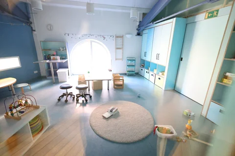 Ein heller, moderner Spiel- oder Klassenraum mit hellblauen Akzenten, einem großen Bogenfenster, Kindermöbeln und Spielzeug.