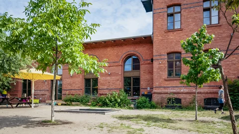 Ziegelgebäude mit Bäumen und Außengelände, inklusive gelbem Sonnenschirm, Picknicktisch und einem Sandkasten.