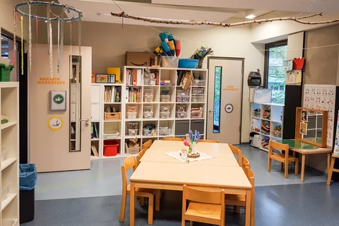 Heller Bastelraum mit Holztischen und Stühlen für Kinder, vollgestellten Regalen, Türen zu Kreativ- und Lego-Werkstätten.