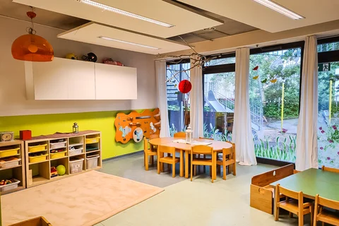 Ein farbenfroher Kindergartenraum mit Kindertischen, Stühlen, Stauraum und Blick auf einen Spielplatz.