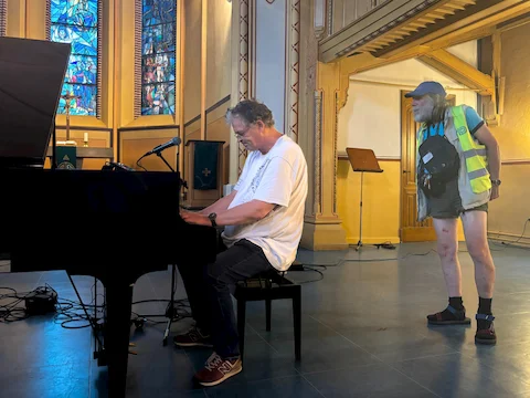 Ein Mann sitzt an einem schwarzen Flügel und spielt, während ein anderer Mann mit grauem Bart, langer Haaren, gelber Warnweste und kurzen Hosen daneben steht und zuschaut. Im Hintergrund sind hohe, verzierte Wände mit goldenen Elementen und bunte Glasfenster zu sehen. Die Szene spielt in einem Kirchenraum mit hellen, warmen Farben.