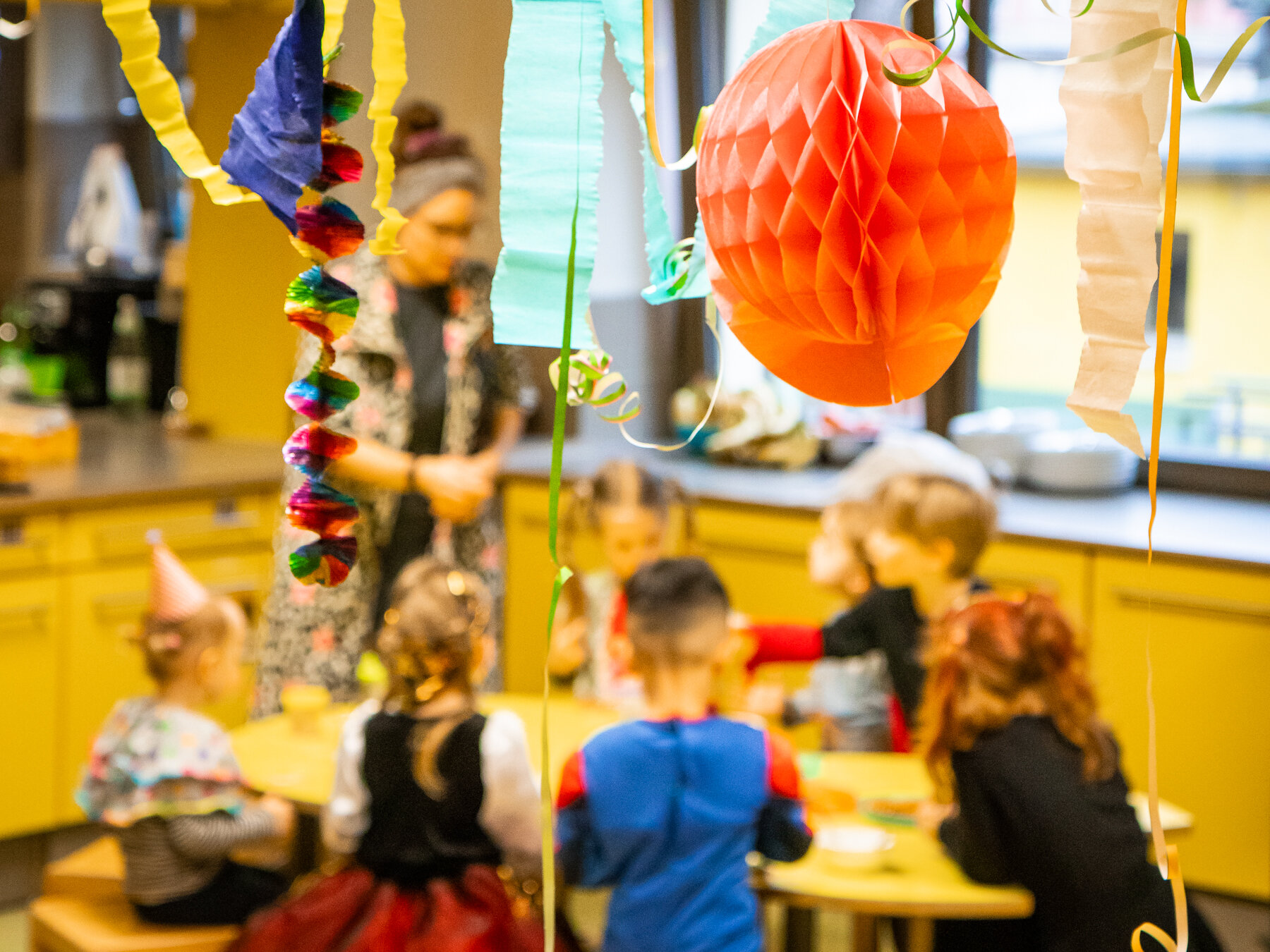Festliche Kinderparty mit Kindern um einen gelben Tisch, einem Erwachsenen im Hintergrund und bunten Papierdekorationen.