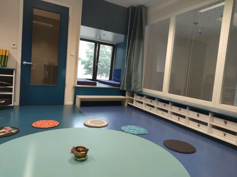 Heller Kinderraum mit einem großen hellblauen runden Teppich, Klangschale, Fenstersitz mit Kissen und Ablageregalen.