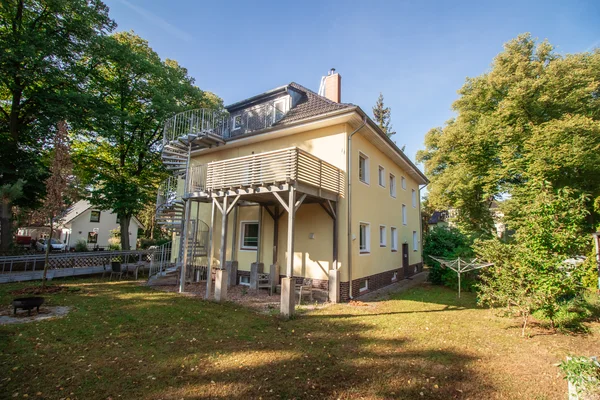 Gelbes Haus mit Balkon, externer Wendeltreppe, und Garten mit Bäumen und Feuerstelle an einem sonnigen Tag.