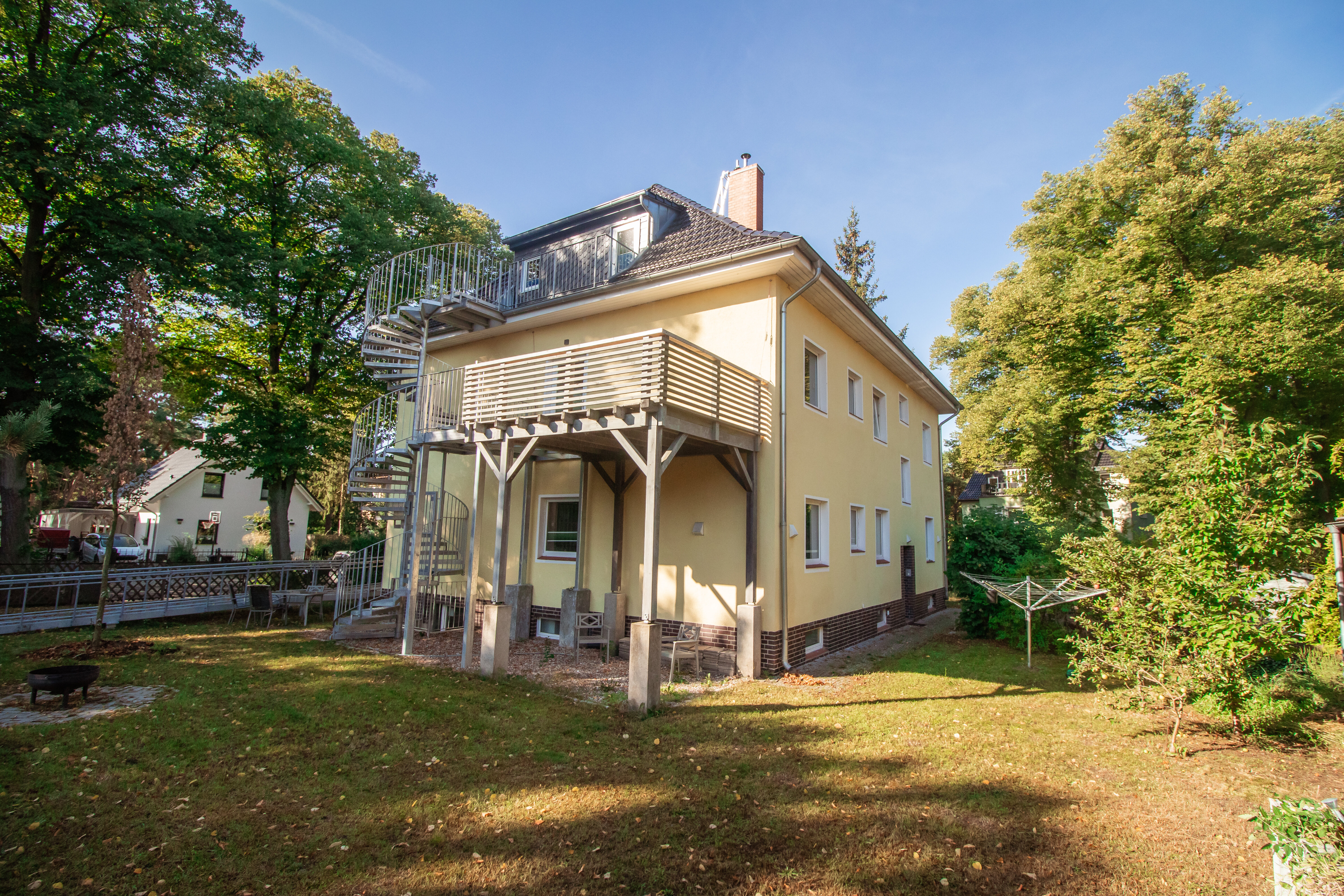 Gelbes Haus mit Balkon, externer Wendeltreppe, und Garten mit Bäumen und Feuerstelle an einem sonnigen Tag.