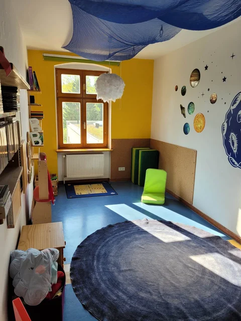 Buntes Kinderzimmer mit Weltraumdeko, blauem Stoff an der Decke, Bücherregalen, Fenster und Sitzkissen.