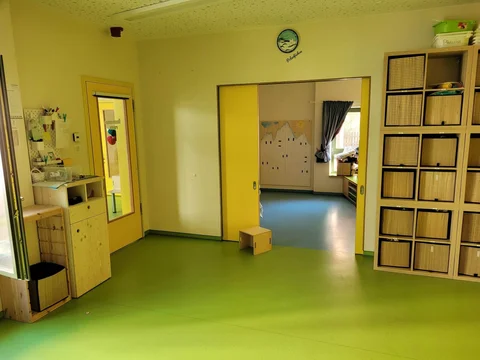 Heller, farbenfroher Klassenraum mit grünem Boden, gelben Wänden, Holzregalen und Blick in einen angrenzenden Raum.