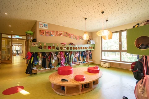 Heller, freundlicher Kindergartenraum mit Garderobe, Kinderkleidung und Schuhen, einer ovalen Holzinsel und Fenstern.