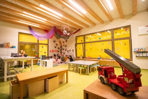 Ein heller, moderner Kindergartenraum mit Kindern auf einer Matte, Tischen und einem großen Spielzeug-Feuerwehrauto vorne.