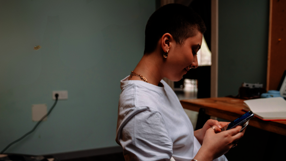 Eine Person sitzt in einem Zimmer. Es gibt ein Bett und einen Tisch. Die Person hat kurze Haare und Piercings. Sie schaut auf ihr Smartphone.