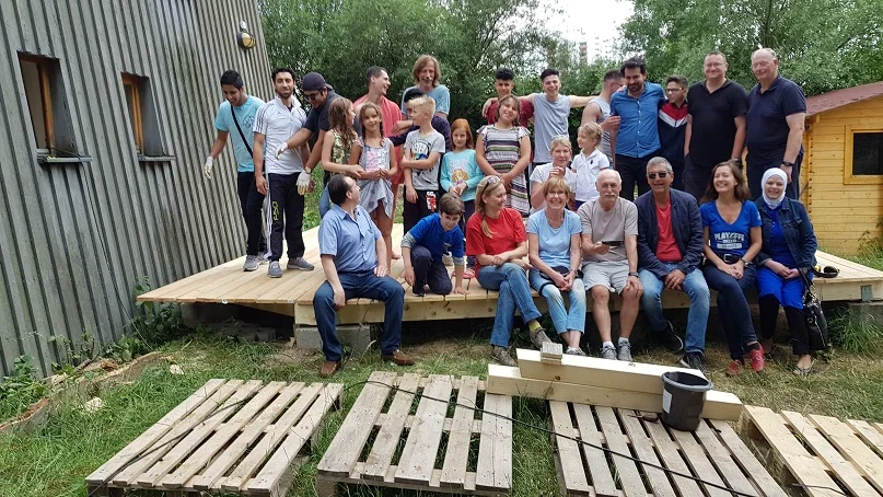 Eine vielfältige Gruppe von Erwachsenen und Kindern lächelt auf einer neu gebauten Holzterrasse, mit Baumaterialien auf dem Gras.