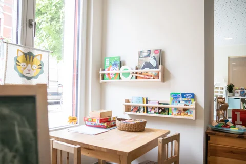 Gemütlicher Lesebereich für Kinder mit Tisch, Stühlen und zwei Wandregalen voller bunter Kinderbücher und Spielen.