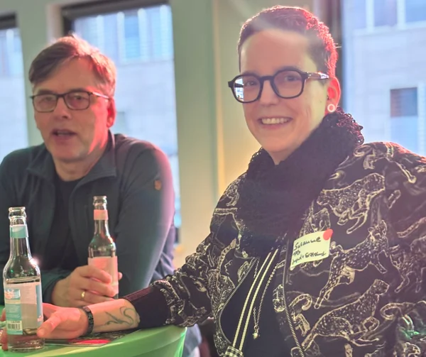 Zwei menschen an einem Stehtisch lächeln in die Kamera in der Hand je ein Getränk. 