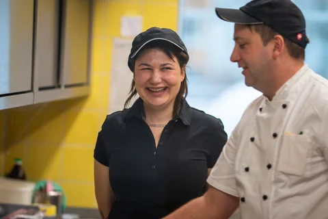 In der Inklusionsküche der SozDia Stiftung Berlin stehen zwei Personen. Eine Frau und Ein Mann. Sie lacht und hat dunkle halblange Haare. Er trägt eine Kochschürze.