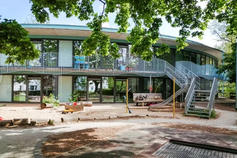 Moderner Kindergarten mit geschwungenem Gebäude, Sandspielplatz, Rutsche und Trampolin unter grünen Bäumen an einem sonnigen Tag.
