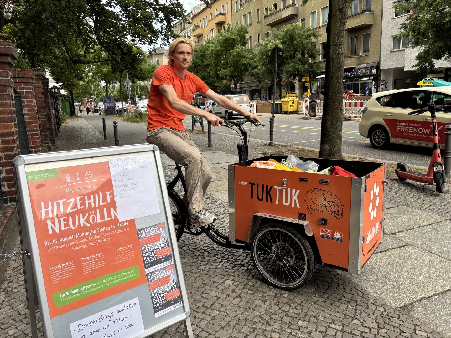 Ein Mann mit neutralem Gesichtsausdruck sitzt auf einem orangefarbenen Lastenrad mit der Aufschrift 'TUKTUK' neben einem Aufsteller mit dem Text 'HITZEHILFE NEUKÖLLN' auf einem Gehweg