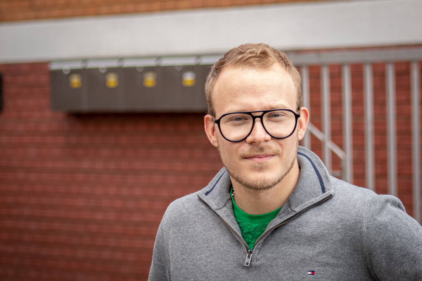 Porträt eines lächelnden Mannes mit kurzen braunen Haaren, Brille, grünem Shirt und grauem Pullover vor Ziegelwand.