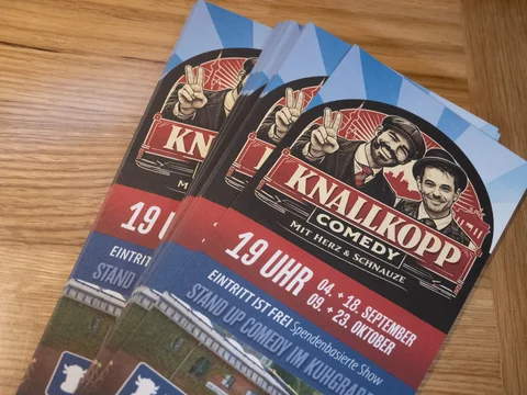 Zwei Flyer für die Stand-up-Comedy-Veranstaltung "Knallkopp Comedy" mit zwei Männern, die jeweils ein Peace-Zeichen zeigen, auf einem Holztisch