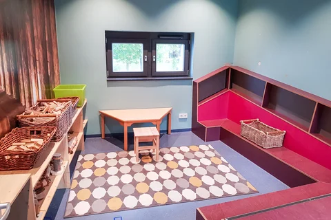 Heller Spielbereich mit mintgrünen Wänden, Holztisch, gemustertem Teppich, roten Sitzelementen und Regalen mit Spielmaterialien.