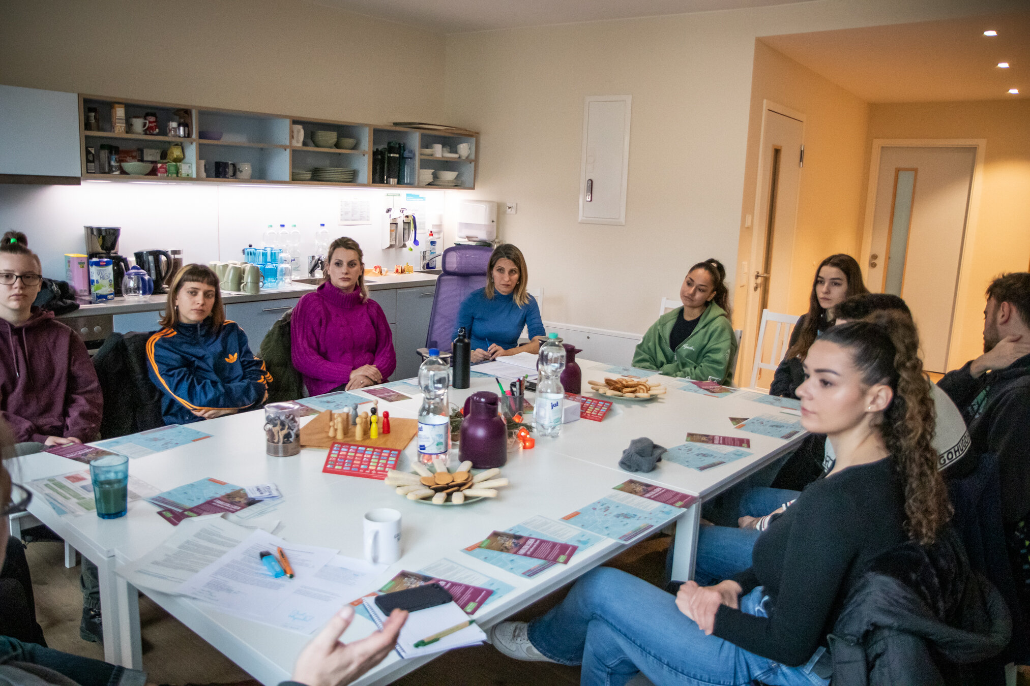 Eine Gruppe junger Erwachsener sitzt an einem Tisch in einem hellen Raum und blickt nach vorne, möglicherweise in einer Besprechung.