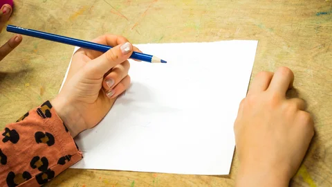Kinderhände halten einen blauen Buntstift und schreiben auf weißem Papier auf einem Holztisch.