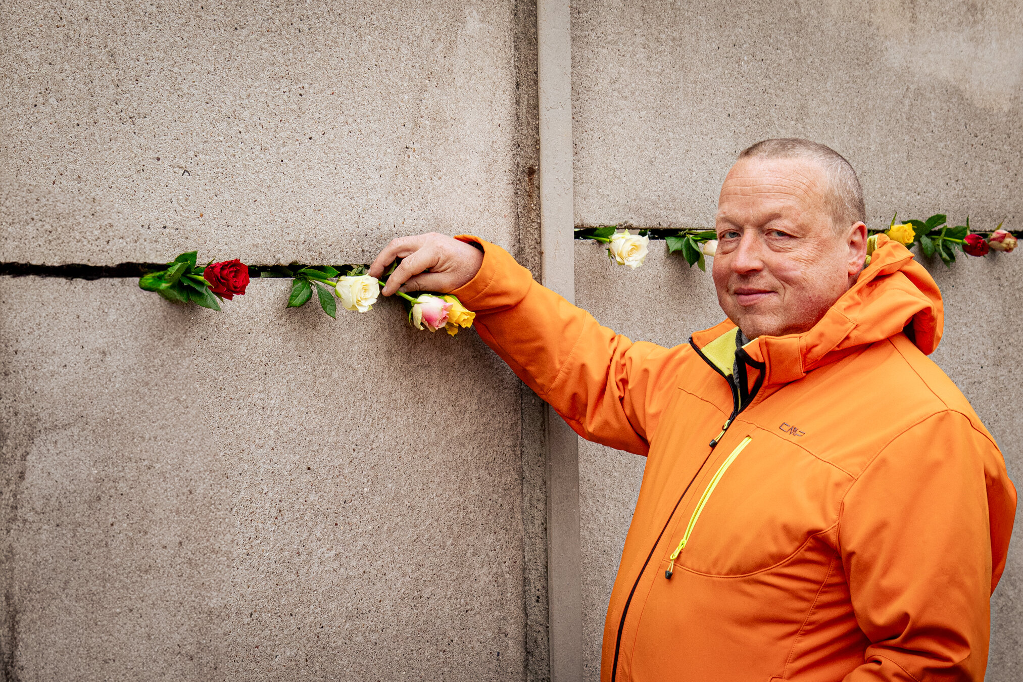 Ein Mann in orangefarbener Jacke lächelt und steckt eine Rose in einen Riss in einer Betonwand mit weiteren Rosen.
