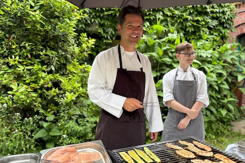 Zwei Personen in Kochkleidung stehen an einem Grill mit verschiedenen Fleisch- und Gemüsestücken, im Hintergrund grüne Pflanzen und eine Backsteinmauer