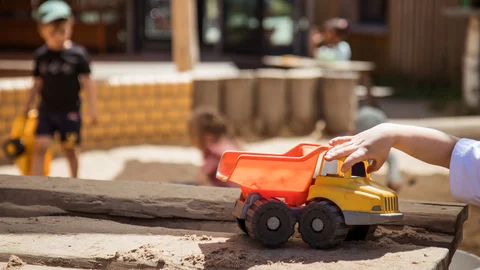Eine Kinderhand spielt mit einem orange-gelben Spielzeugkipper im Sandkasten, im Hintergrund weitere Kinder.