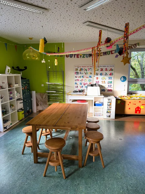 Innenraum mit großem Tisch, Sitzhockern und Regalen mit Spiel- und Bastelmaterial.
