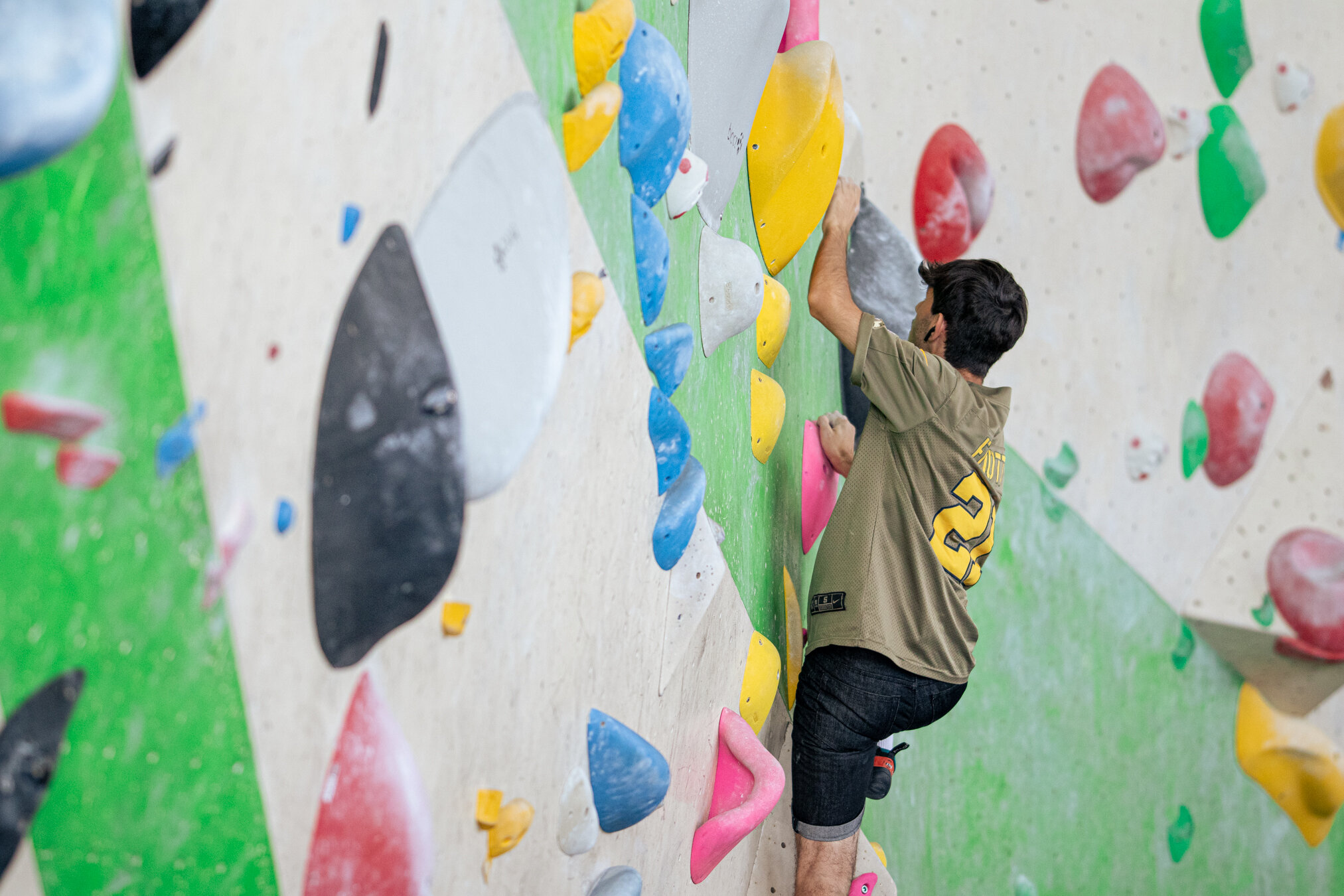 Mann bouldert an einer bunten Kletterwand, greift nach einem gelben Griff.