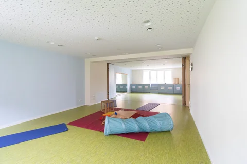 Geräumiger Raum mit hellgrünem Boden, hellblauen und weißen Wänden sowie Kinderspielgeräten wie einem blauen Tunnel und Matten.