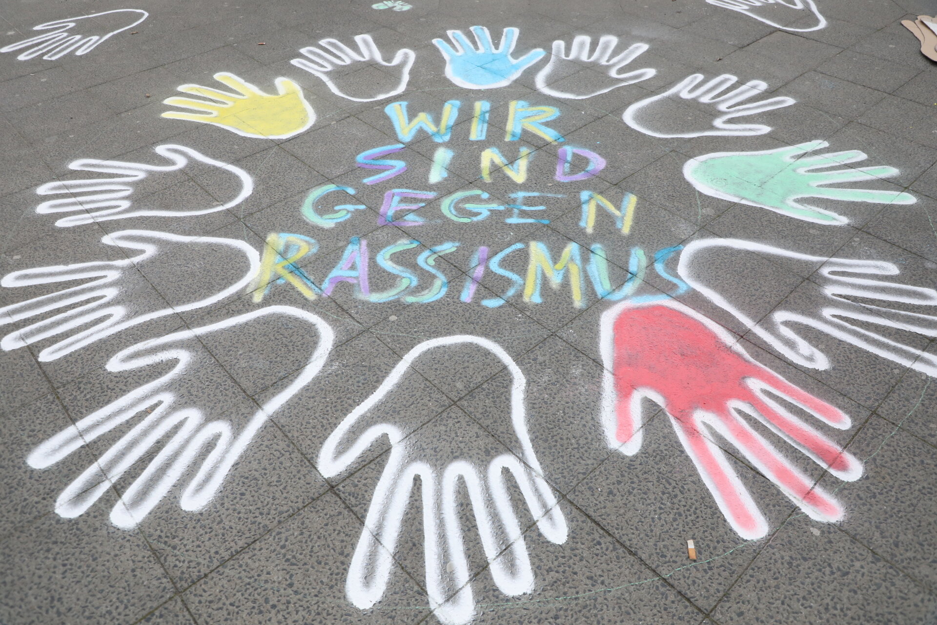 Kreidezeichnung auf dem Boden: Umrisse von Händen in einem Kreis, mit dem Text "WIR SIND GEGEN RASSISMUS" in der Mitte.
