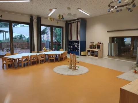 Heller, geräumiger Kindergartenraum mit langem Tisch, kleinen Stühlen, Spielzeugregalen und großen Fenstern zu einer Terrasse.