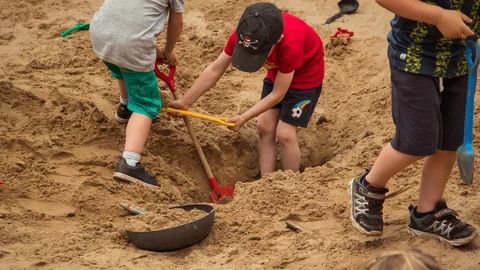 Drei Kinder spielen in einem großen Sandkasten und graben aktiv mit roten, gelben und blauen Schaufeln.
