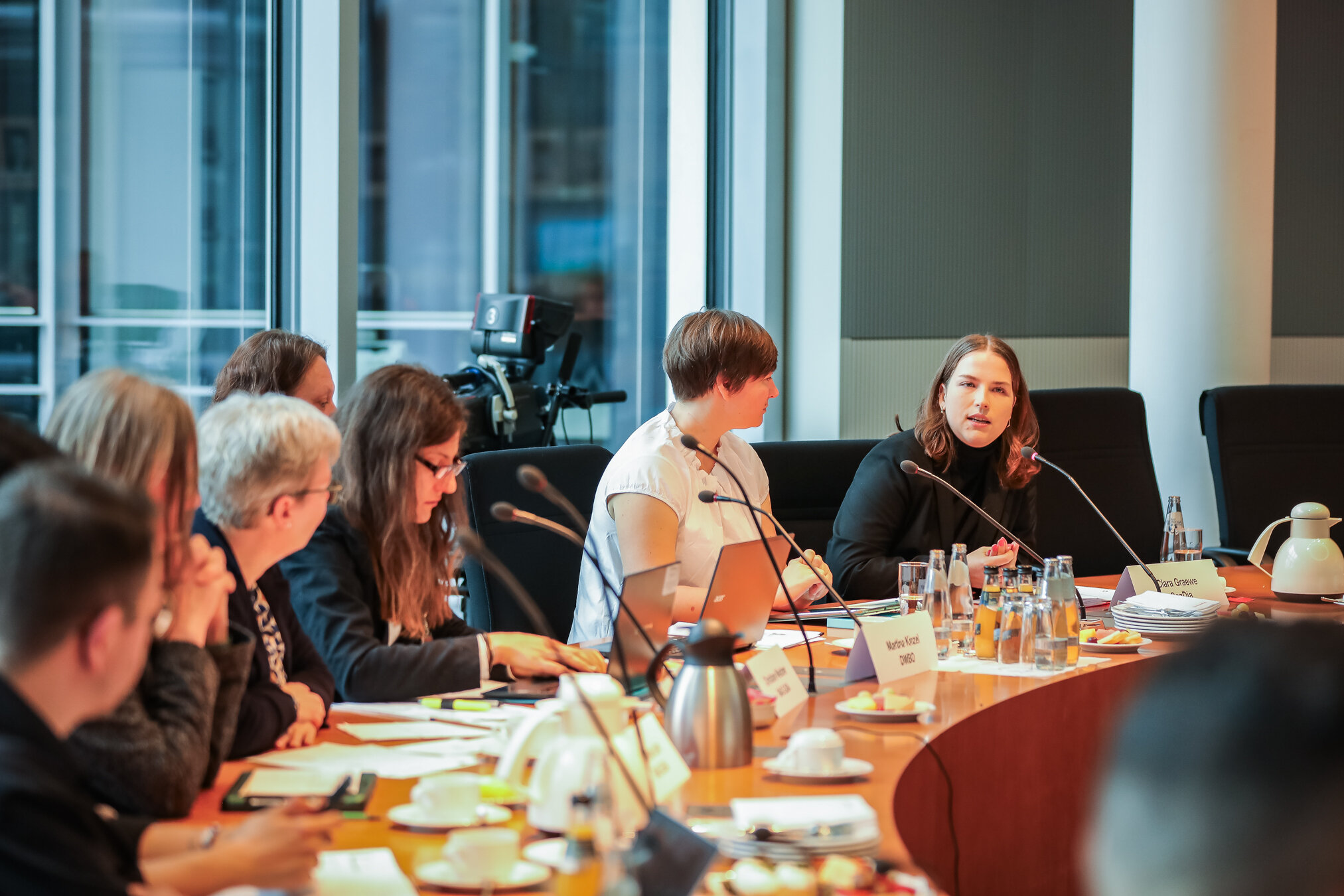 Eine Frau spricht in ein Mikrofon an einem Konferenztisch, umgeben von verschiedenen Teilnehmern, Laptops und Erfrischungen.