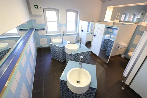 Heller, farbenfroher moderner Waschraum mit mehreren Waschbecken auf blauen Mosaik-Säulen, blau-weiß gefliesten Wänden und Toilettenkabinen.