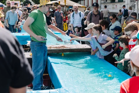 Mann mit gelbem Hut und grünem T-Shirt steht an einem Wasserbecken und reicht Kindern mit Kescher kleine gelbe Enten