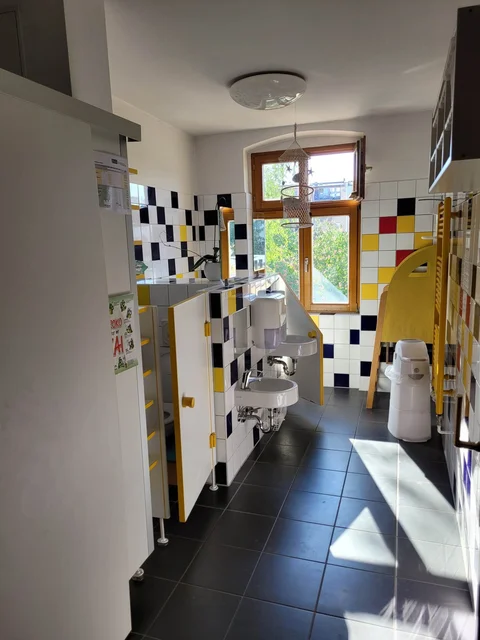 Helles, farbenfrohes Kinderbad mit kleinen Waschbecken, Schachbrettfliesen, kurzen WC-Kabinen, Fenster und Sonnenlicht.