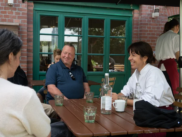 Zwei Personen sitzen an einem Holztisch im Freien und unterhalten sich, auf dem Tisch stehen Gläser, eine Wasserflasche und eine Kaffeetasse