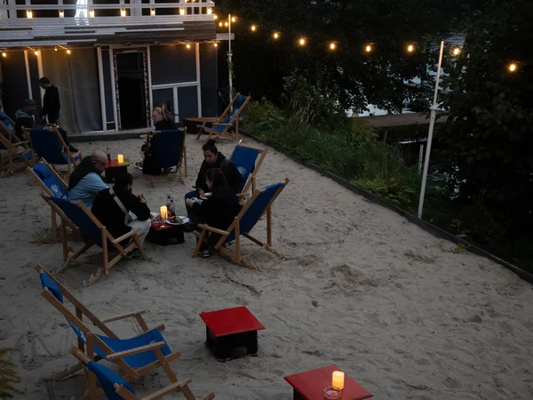 Mehrere Personen sitzen abends auf blauen Klappstühlen im Sand vor einem Haus mit Lichterkette und Kerzen auf kleinen roten Tischen